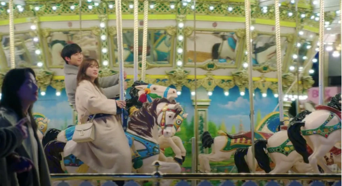 A man and a woman sitting on a carousel with a woman off to the side in shadow