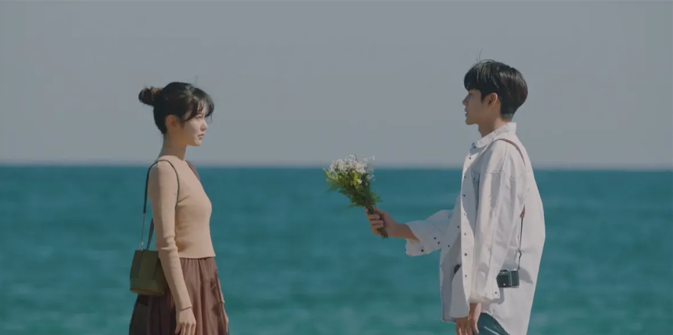 A man offering a woman a bouquet of white flowers by turquoise water
