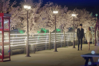 A man and woman walking along a lamplit trail with cherry trees in the background