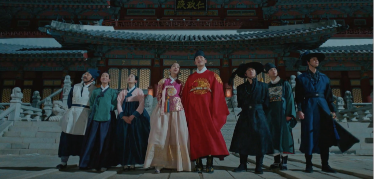 Three women and five men in hanboks standing in front of a palace at dusk