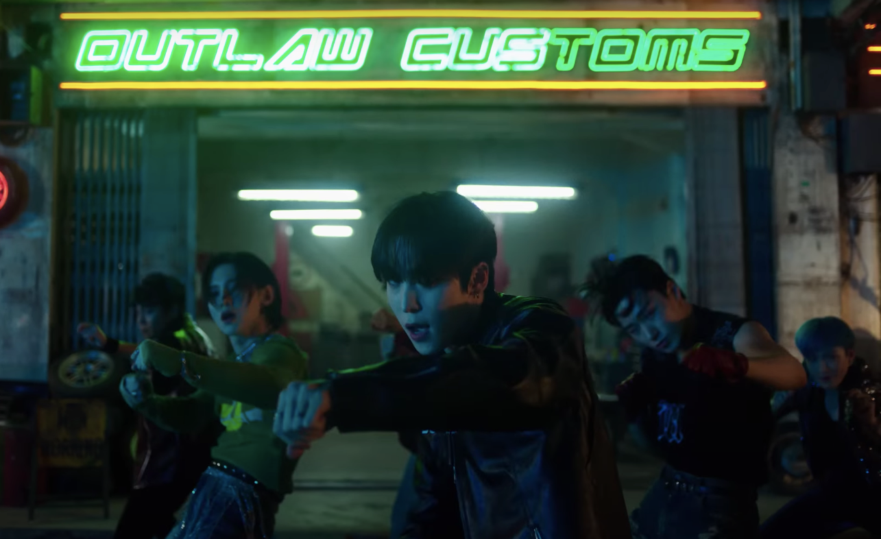 Eight men dancing in front of neon green "Outlaw Customs" sign