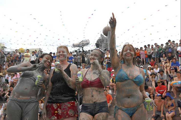 Four women in swimsuits covered in mud standing in front of a muddy crowd