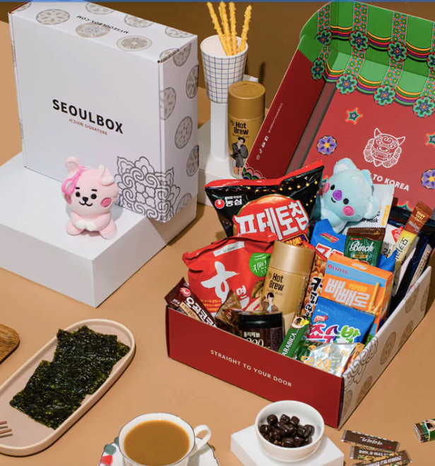An opened Seoulbox beside two unopened boxes with mugs of coffee and a pink BT21 plushie