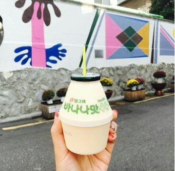 A person's hand holding a banana milk cup in front of a geometric mural in an alley