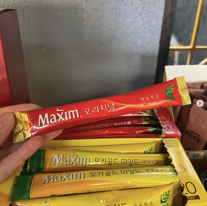 A person's hand holding a Maxim instant coffee packet above a box with more packets