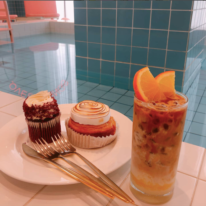A glass of iced coffee with orange slices beside a plate of cakes near pool tiling