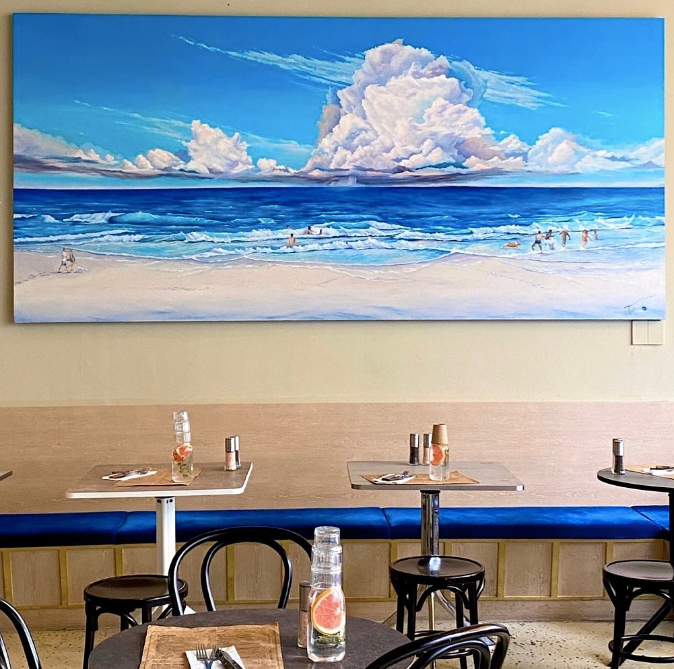 Blue tables and gray chairs under a painting of a beach, ocean, and blue sky