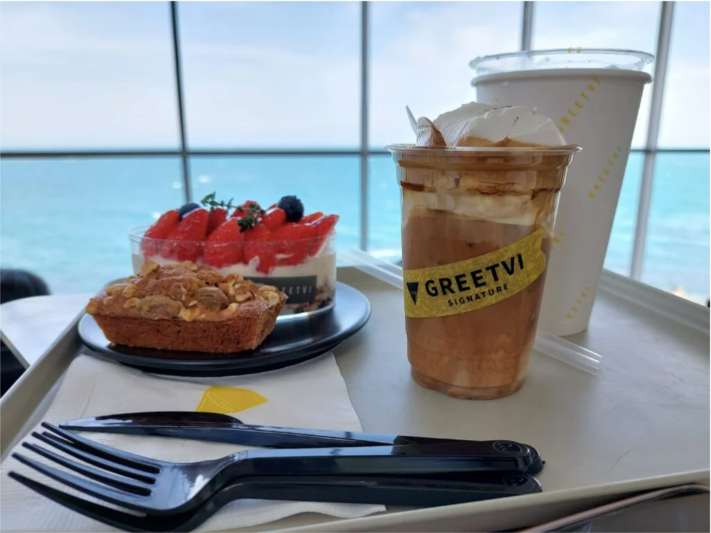 An iced coffee and hot coffee with fruity desserts on a tray with a plastic fork and knife