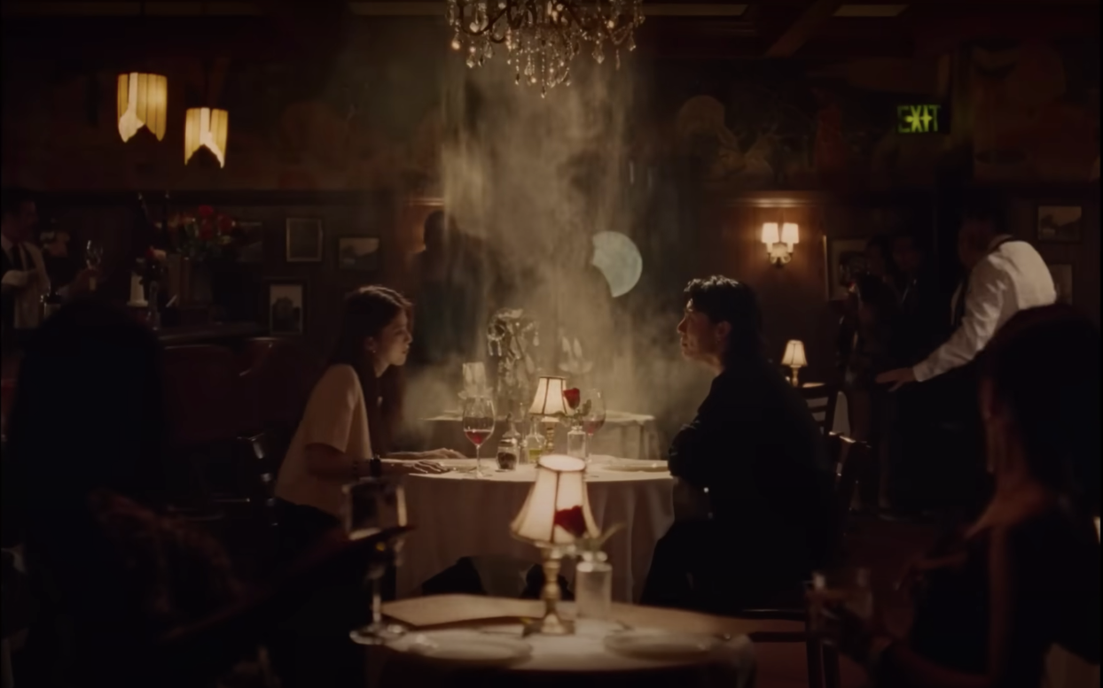 A man and a woman sitting opposite each other at a dinner table as a chandelier falls behind them