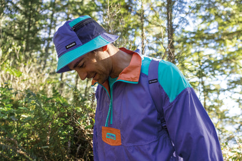 Male model wearing a columbia sportswear company fleece in purple and a matching bucket hat in purple on his head. He is stood in front of bushes in a forest.
