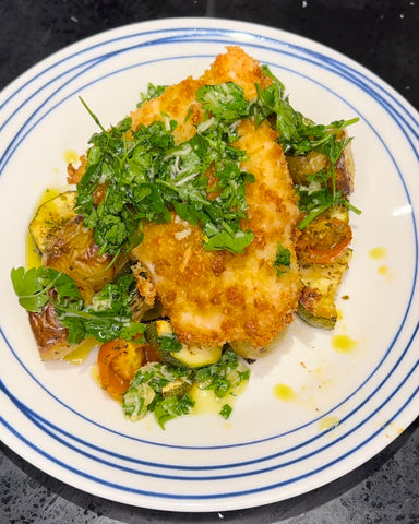 A white plate with panko breadcrumbed chicken placed on top of roasted courgette and cherry tomatoes, dressed with parsley, garlic and olive oil dressing