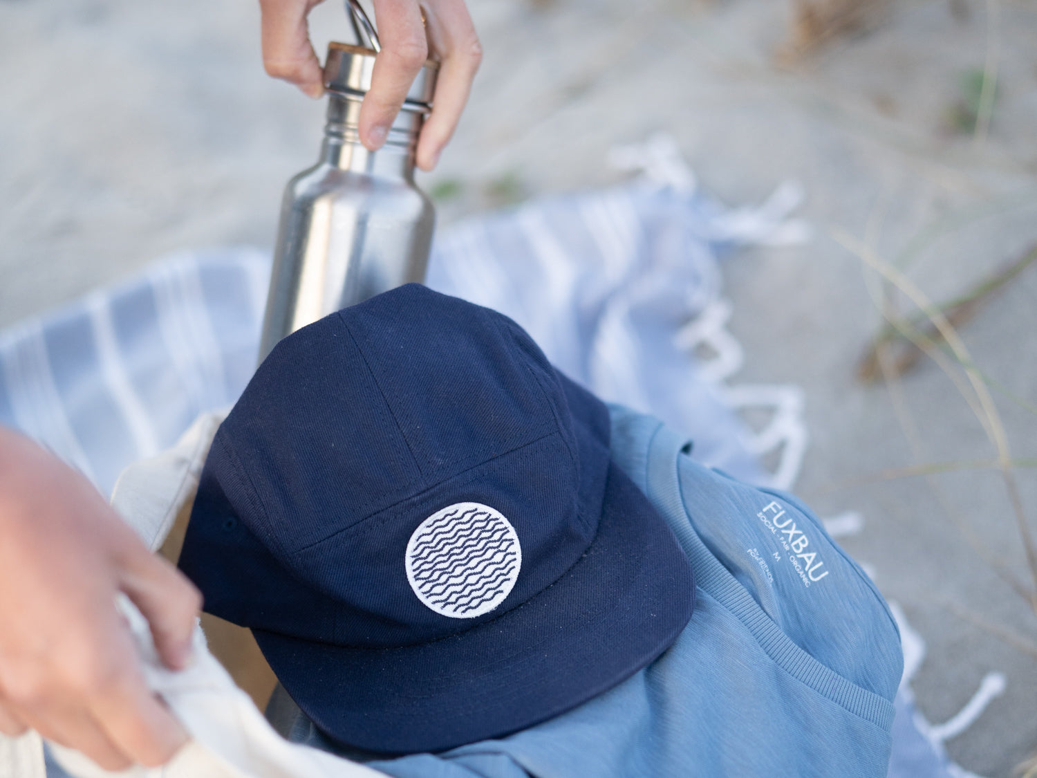 Nachhaltige Essentials für sonnige Strandtage: Trinkflasche, Cap, Badetuch und T-Shirt. 