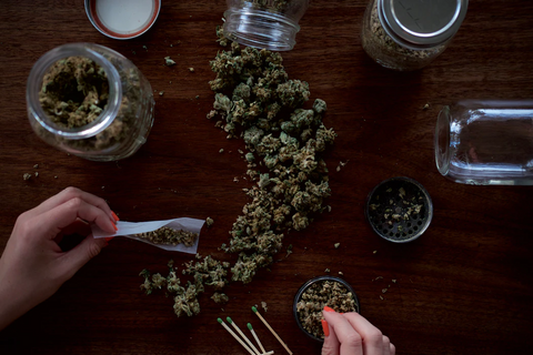 A hand holding a pre-rolled weed cigarette in front of a wooden table with a metal weed grinder  and a glass jar on top of it