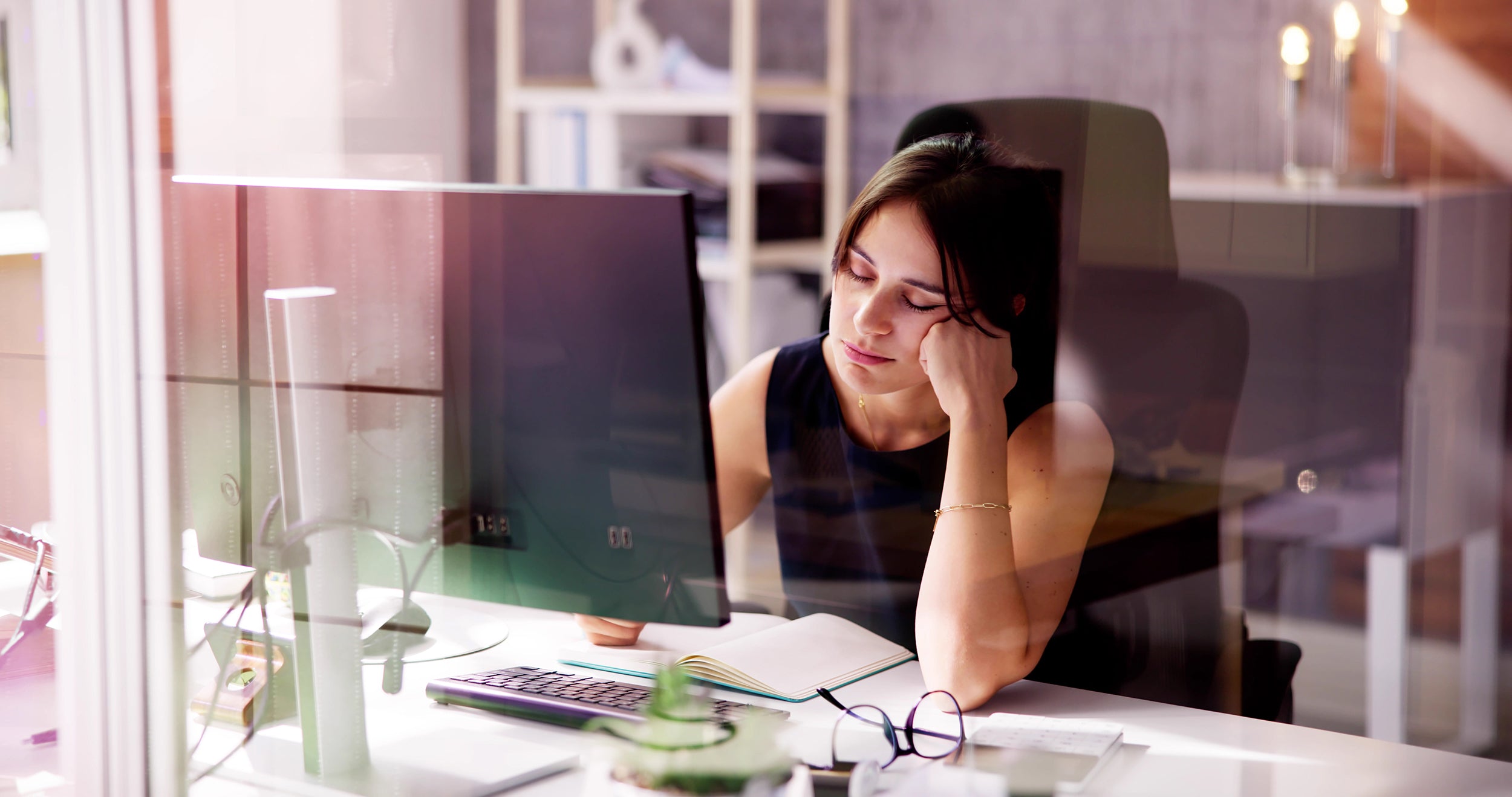 A woman nods off while working at her computer. Learn more about the origins of back pain and the relationship between sleep quality, back pain, and how to get relief in our comprehensive guide.