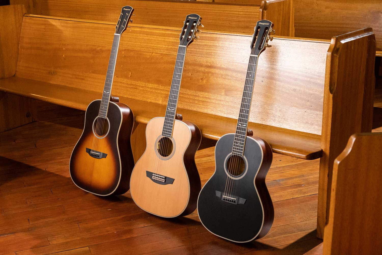 Three guitars lined up against a wooden pew: sloped shoulder, nylon string, and black parlor