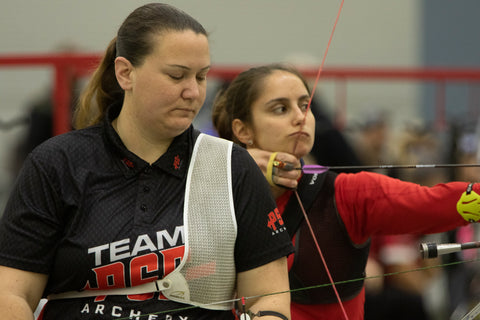 An Olympic recurve archer wearing a PSE jersey waits as the archer behind her shoots an arrow. 