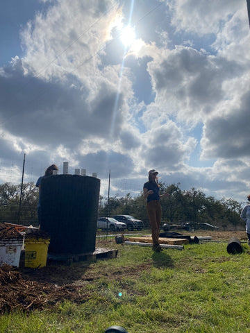Andie Marsh - female Texas soil expert and biologist - talks to a group of mycelium-lovers in the hemp field at Jester King Brewery. Andie is standing in the sunlight next to a four and a half foot tall compost bin she constructed from hardware cloth and pvc pipes.