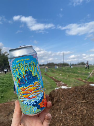 Snorkel Mushroom Oyster beer can, held up by a dirty hand, with a Texas farm field blurred in the background. The beer is made by Jester King Brewery, and has a colorful label of coral and oyster mushrooms, under the water.