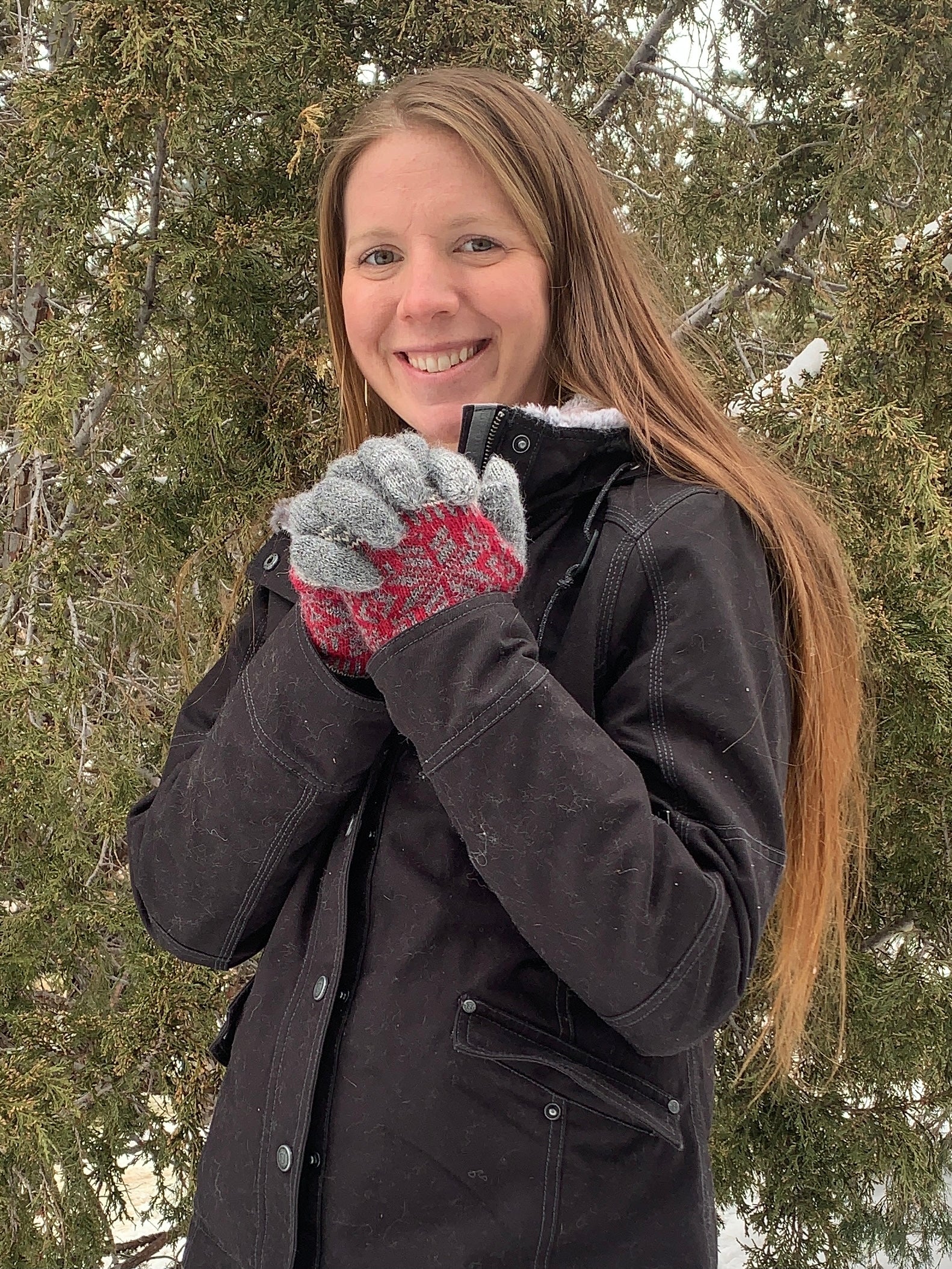 woman smiling, standing in front of a pine tree wearing a black coat and patterned gloves. 