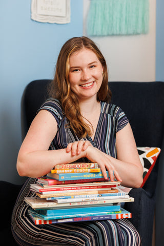 Gina sitting in a navy blue chair, her arms and hands resting on a big stack of books on her lap