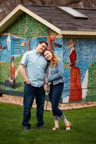 Zach in a light blue shirt standing next to Gina in a denim jacket who has her head on his shoulder. They are standing in front of a small building that is covered in a pointillistic mural.