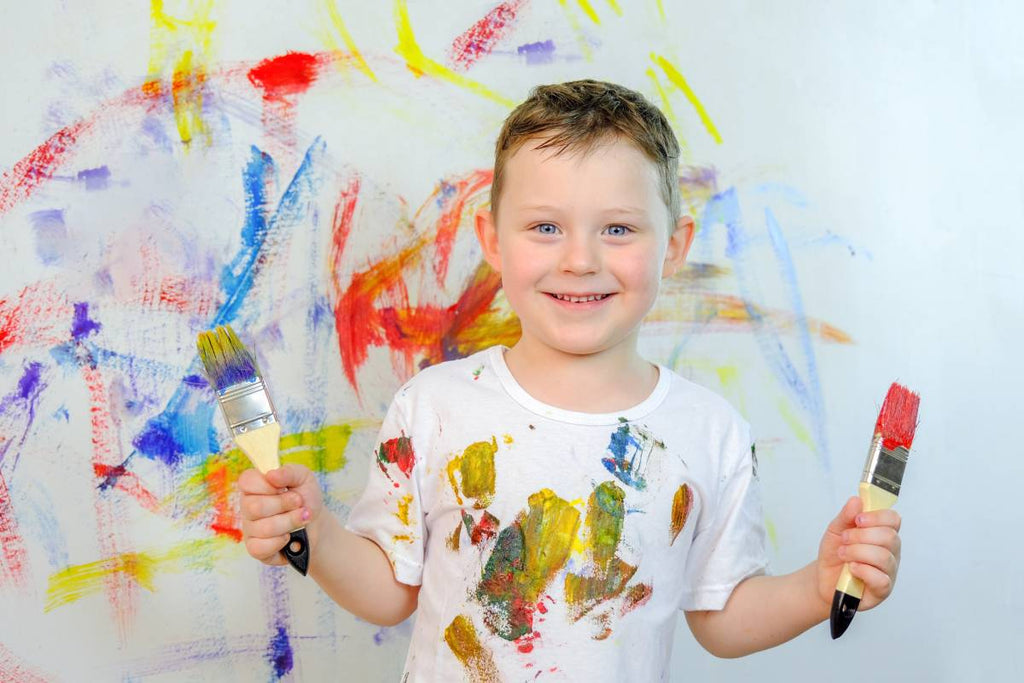 Young boy holding paint brushes wearing shirt covered in acrylic paint