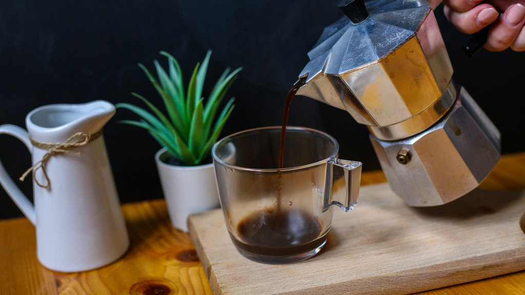 freshly brewed coffee being poured into a clear glass mug from a moka pot