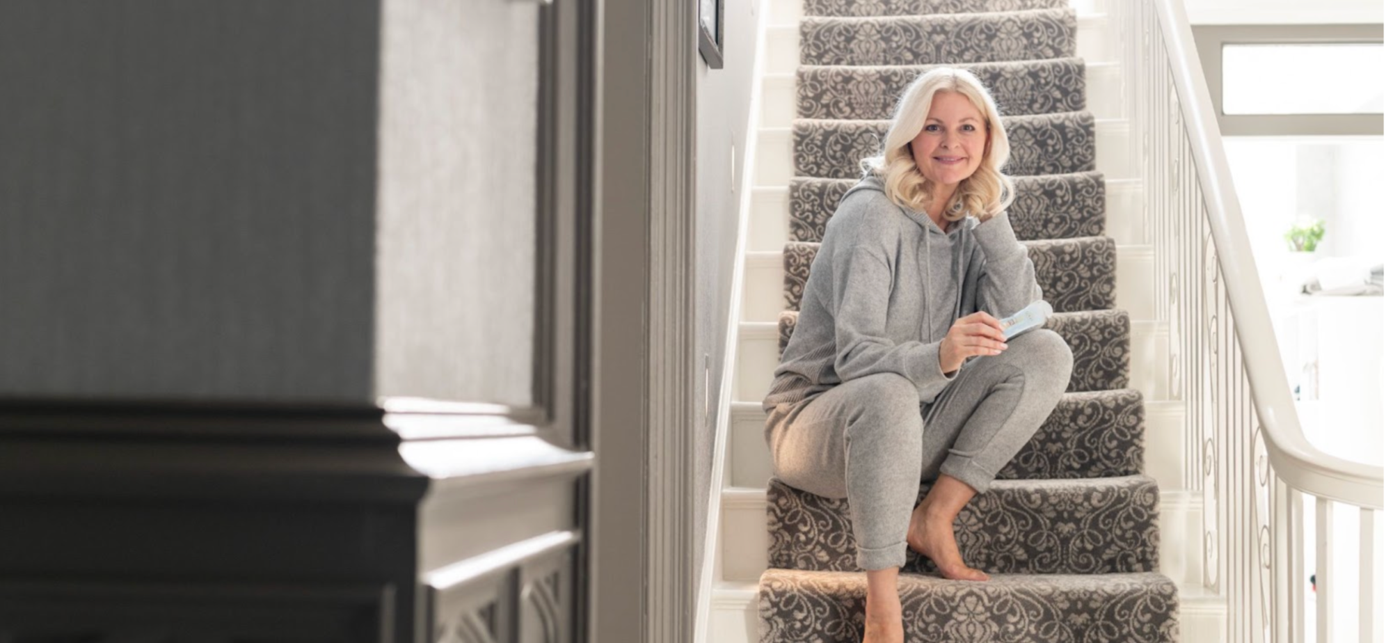 Woman in grey loungewear, sitting on grey stairs, holding an Arella Beauty liquid Collagen Supplement