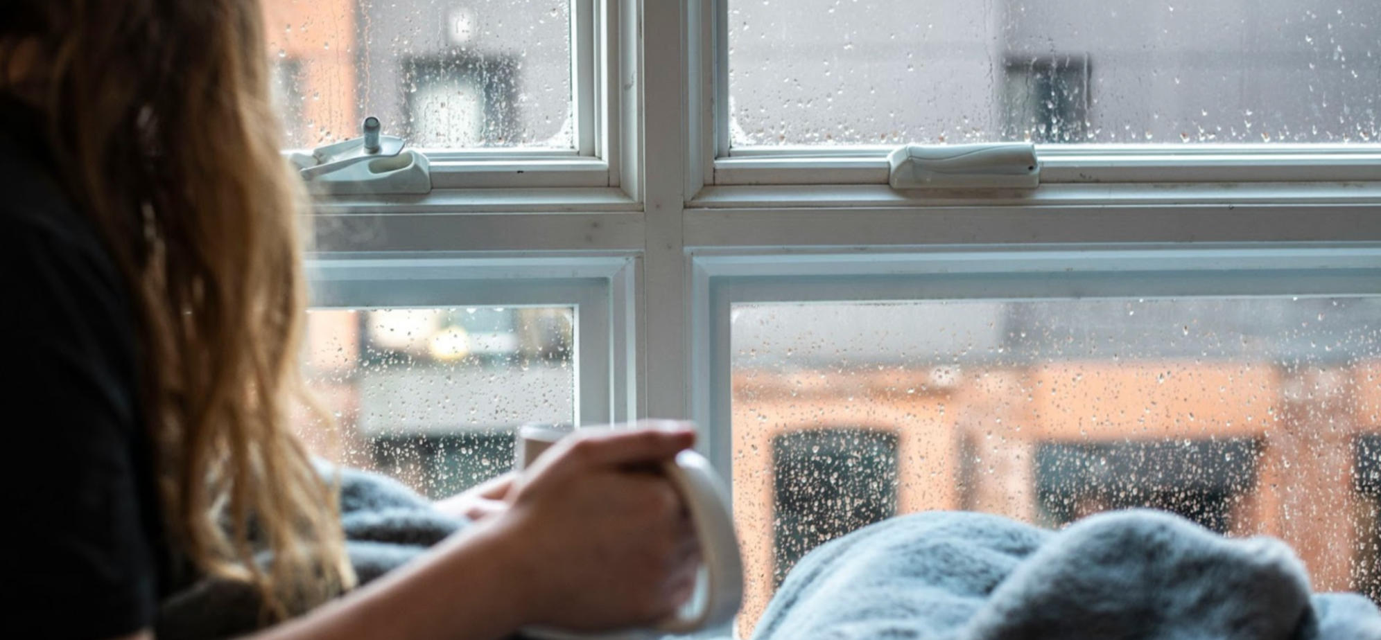 Woman sitting by raining window with a blanket and holding a mug