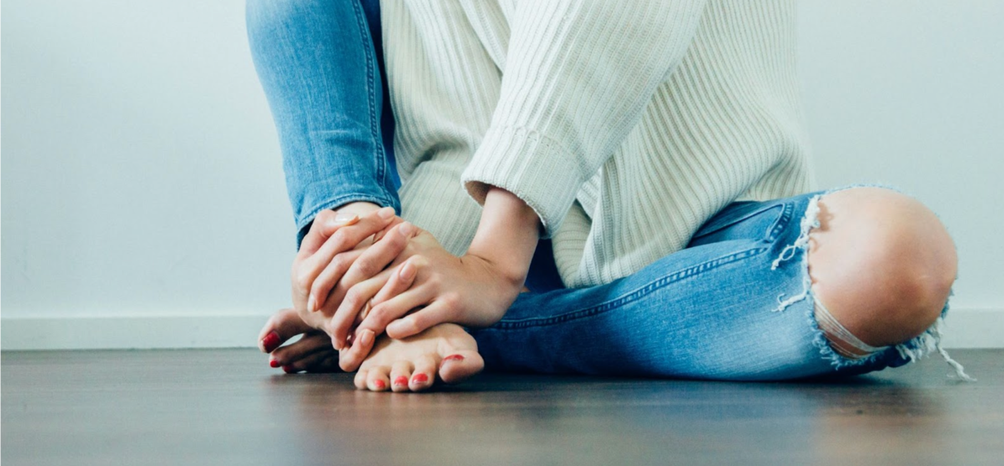 Woman cross-legged on the floor in ripped jeans, holding a sore ankle