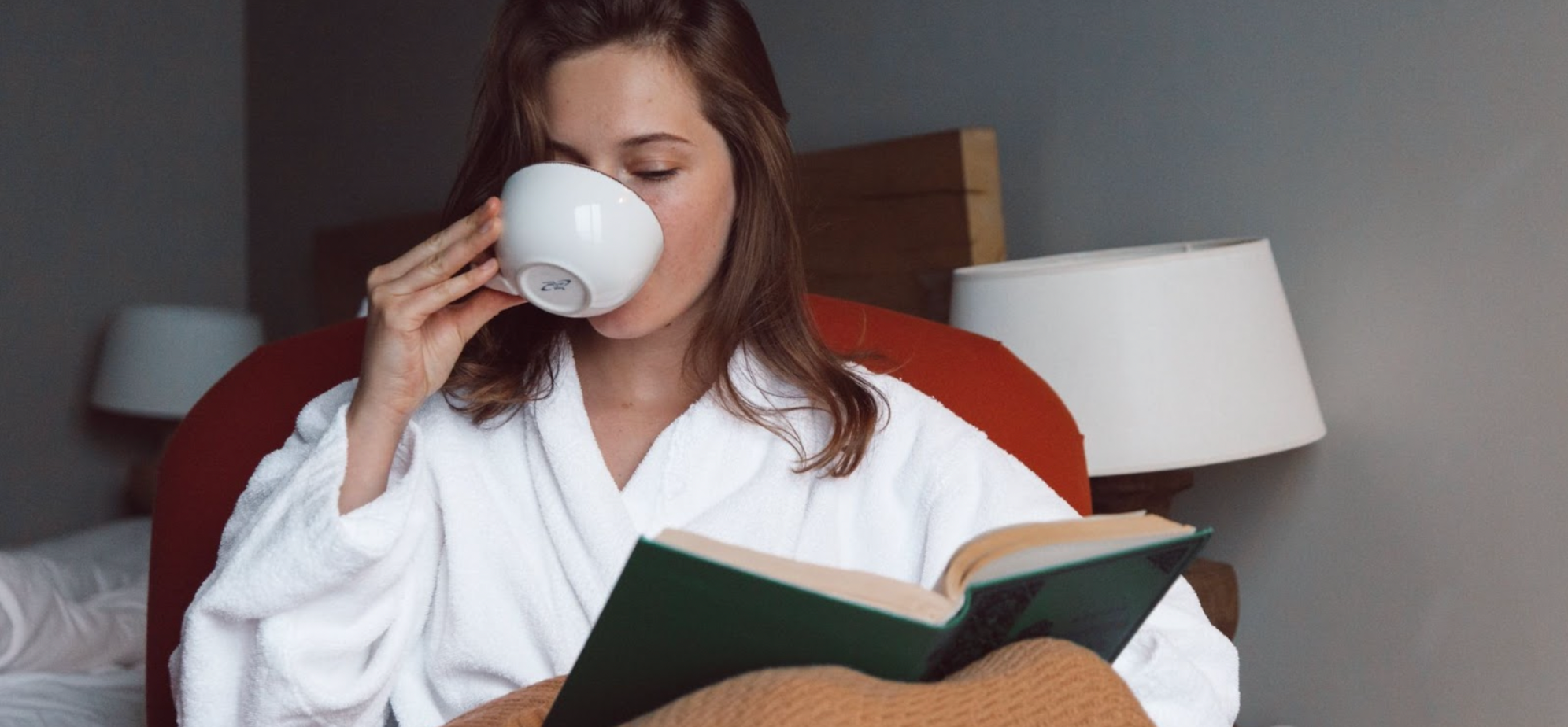 Woman underneath a blanket, sipping a cup of tea and reading a book