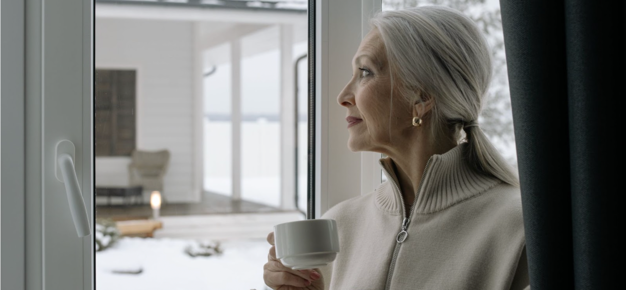 Older woman with grey hair, holding a cup of tea by a window in a sweater