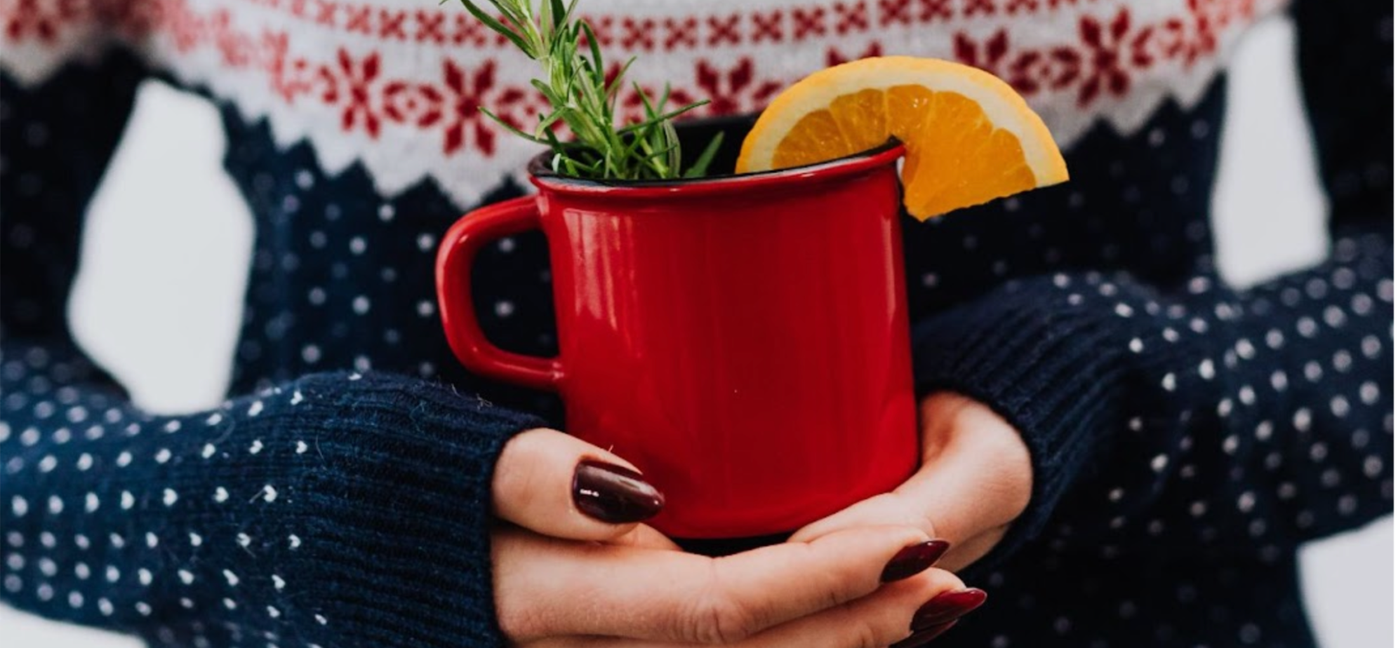 Woman in a Christmas jumper holding a red mug with thyme and an orange slice for a Christmas drink
