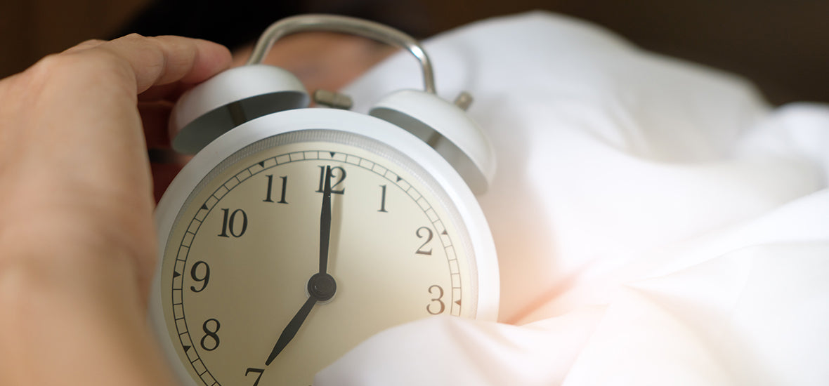 A hand holding white analog alarm clock, sat on top of a white duvet. Man ignoring the alarm because of sleep procrastination.