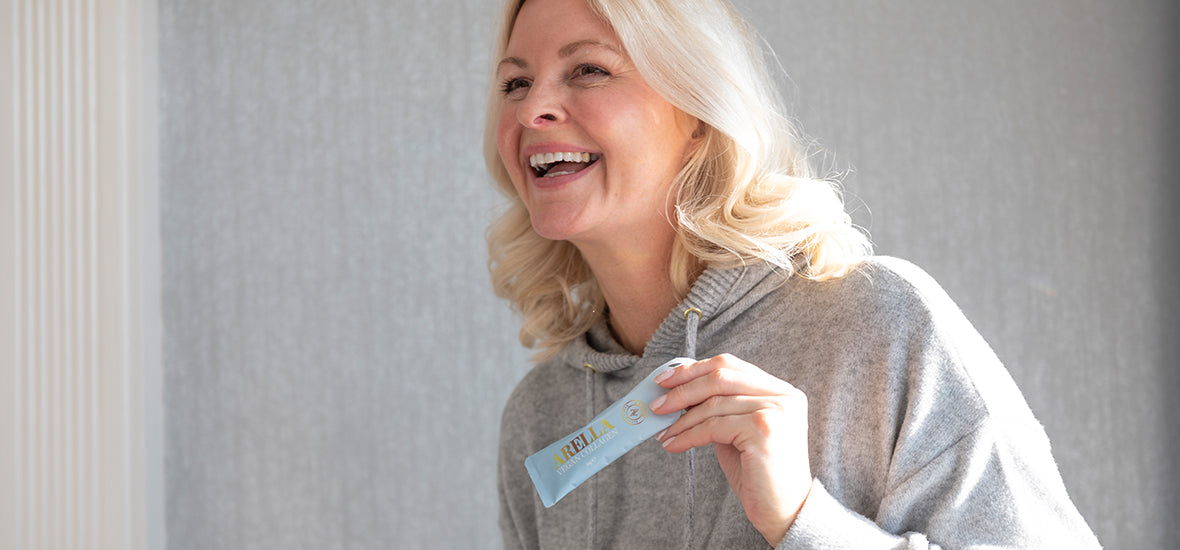 Woman with white blonde hair in a grey hoodie, smiling and holding an Arella Beauty collagen supplement.