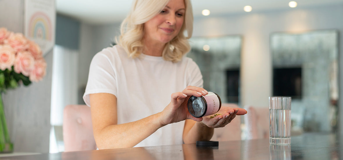 A blonde woman with a white t-shirt sat at a kitchen countertop with a glass of water, tipping Arella Beauty Dream supplements into her hand for her evening routine for sleep.