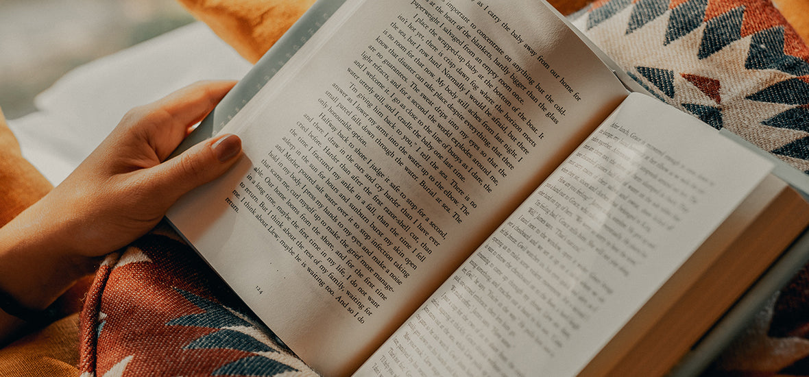 Woman’s hands holding open a book and reading on orange and patterned cushions as part of her evening routine for sleep.