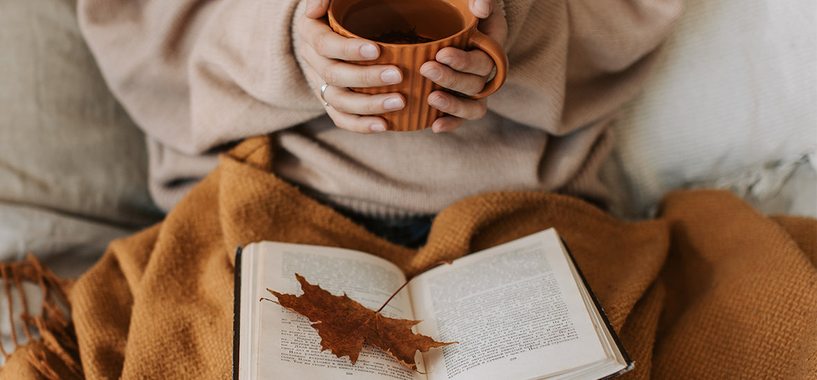 Woman in a beige jumper with a cosy blanket and book, holding a hot drink to keep healthy in autumn.