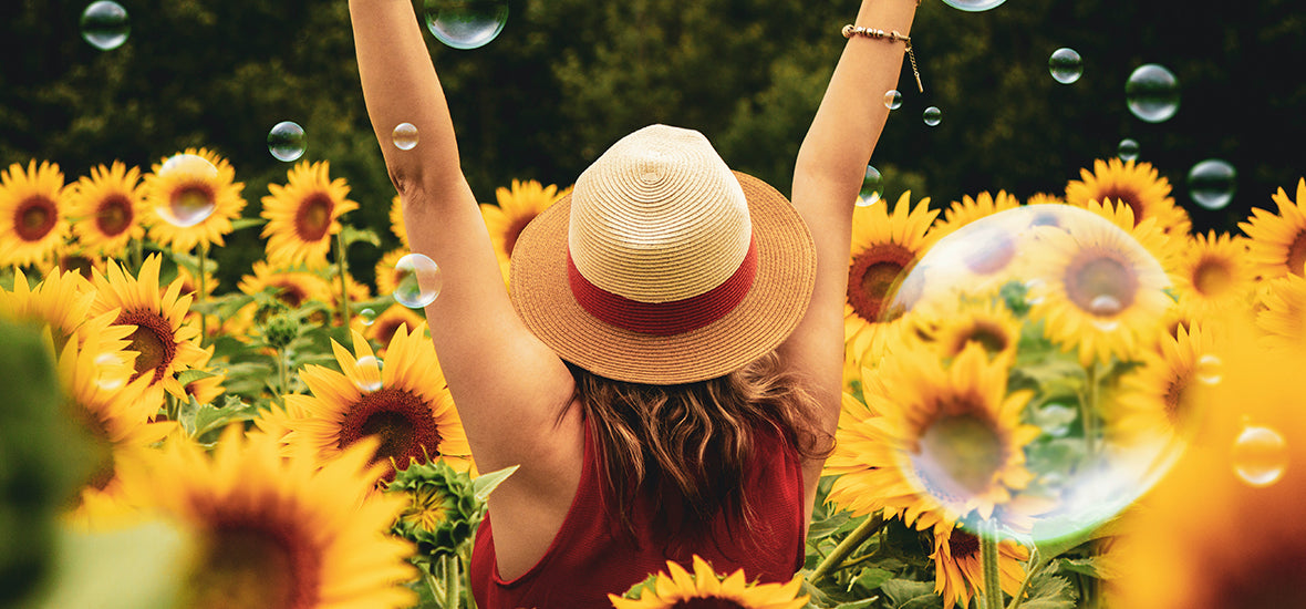 Back of woman wearing a straw hat with her hands in the air, in a field of sunflowers and bubbles.