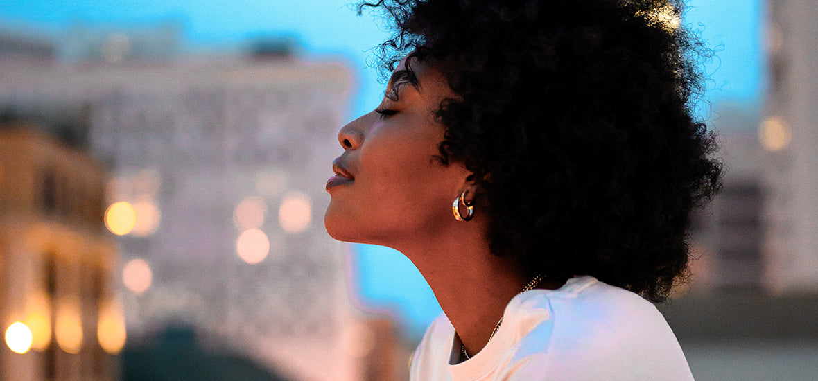 Woman with coily, afro hair and her eyes closed looking over a city and considering when to start taking collagen.