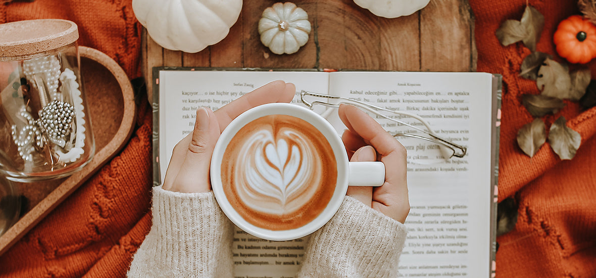 Person holding a latte on a book with pumpkins and a blanket in autumn.