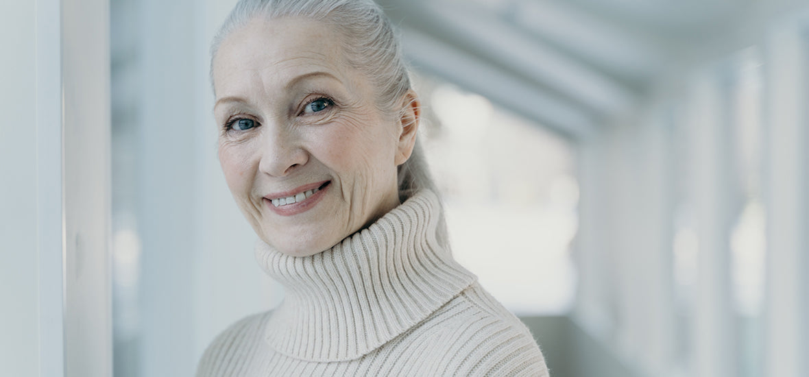 White, older woman with grey hair in a ponytail, wearing a white roll neck jumper and reduced wrinkles from using ginseng for skin.