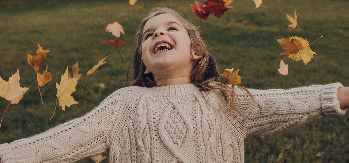 Happy little girl wearing a cosy cream knit jumper, throwing autumn leaves into the air.