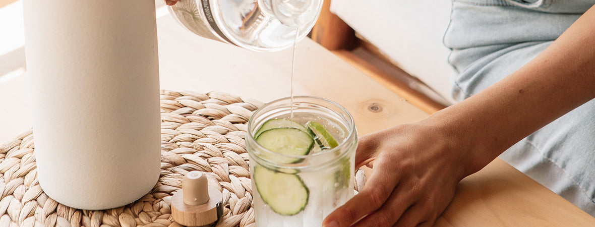 Collagen: Woman pouring a bottle of water into a glass jar of cucumber water.