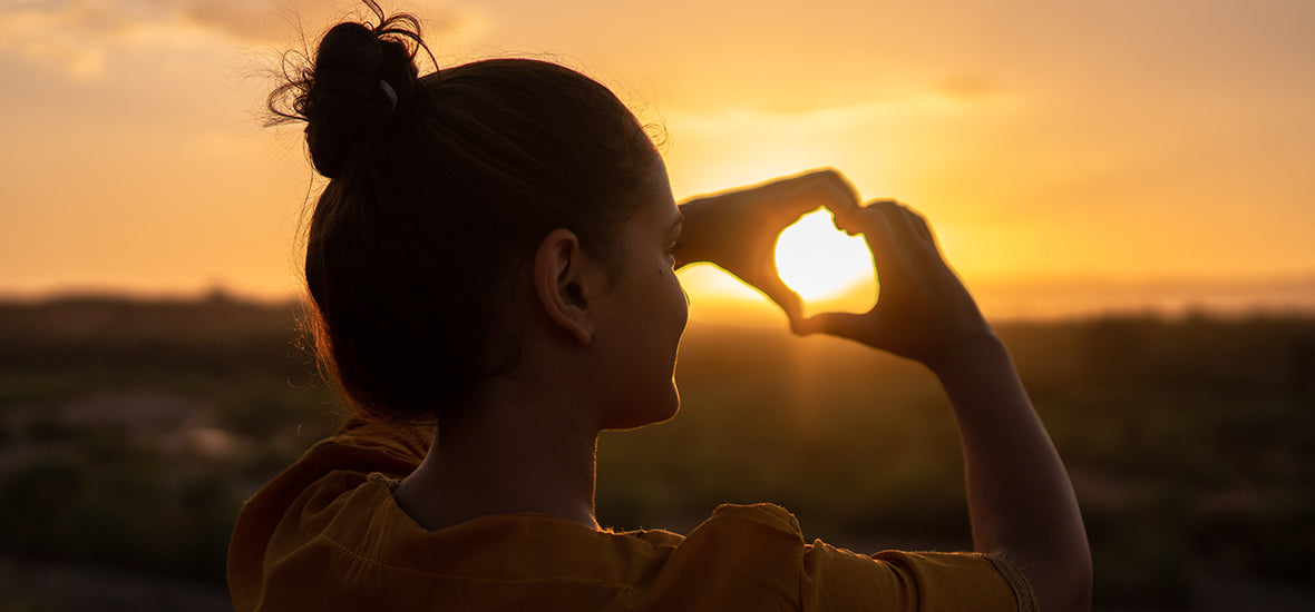 Woman facing the sunset and making a heart with her hands for heart health.