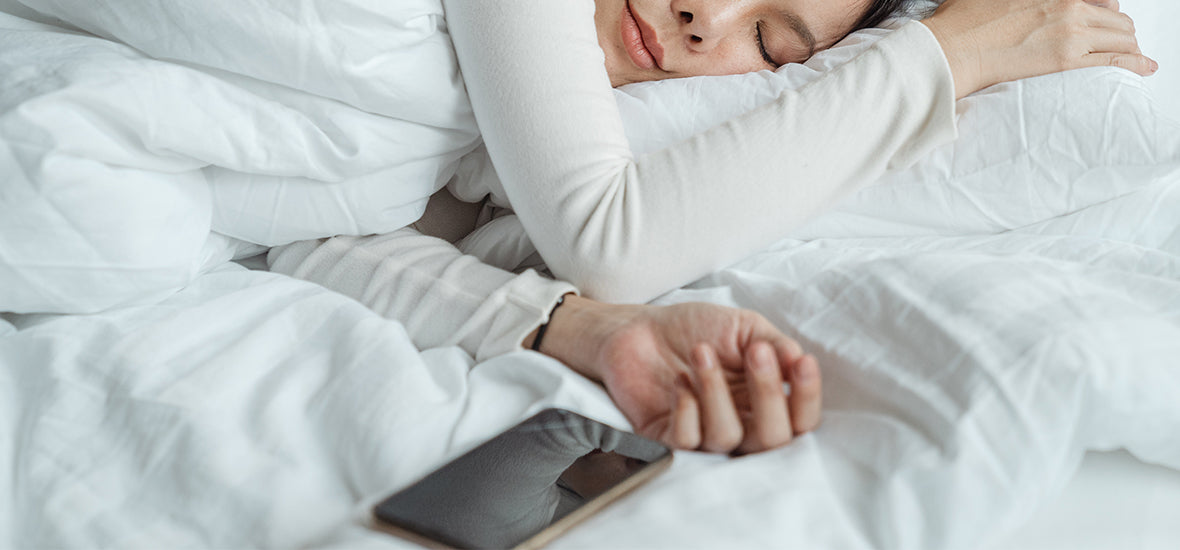 Woman asleep wearing white, under white bedding, with her phone beside her hand after her evening routine for sleep.