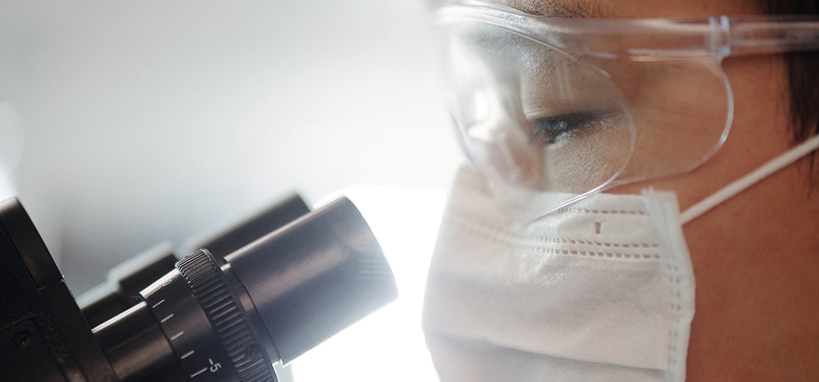 Close-up of a female scientist wearing goggles and a mask looking through lab binoculars.
