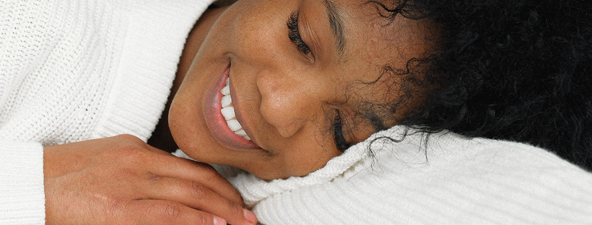 Collagen for gut health: Woman with afro smiling with eyes closed, lying her head on her arm, wearing a white jumper.