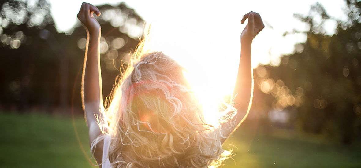 Back of woman's head, with blonde, curly hair, looking at the sun and arms raised to reduce hormonal stress.