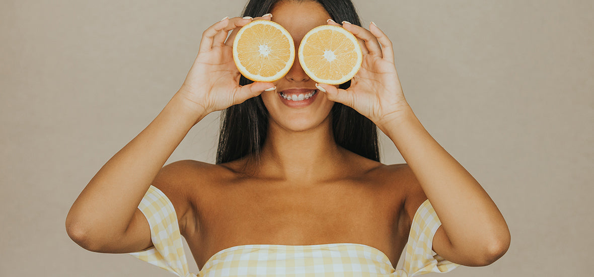 Woman with long dark hair, smiling and holding orange slices in front of her eyes.